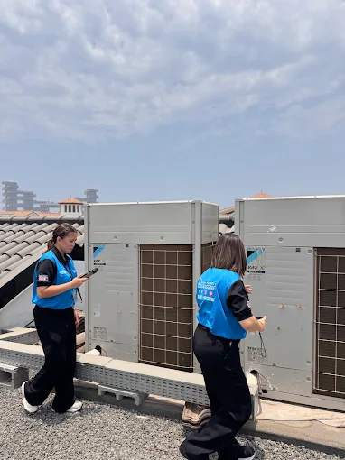 Two technicians in blue safety vests inspect rooftop air conditioning units on a sunny day in an urban setting.