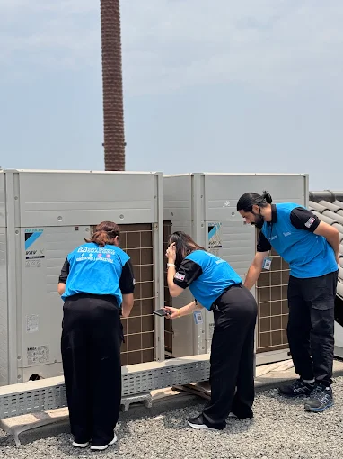 Three technicians in blue uniforms inspect rooftop HVAC units together one pointing at a panel as another leans in with a flashlight on the side of the building