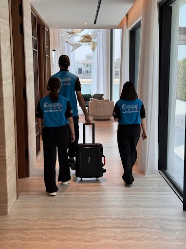 Three staff members in blue uniforms walk down a bright hallway pulling a black rolling suitcase