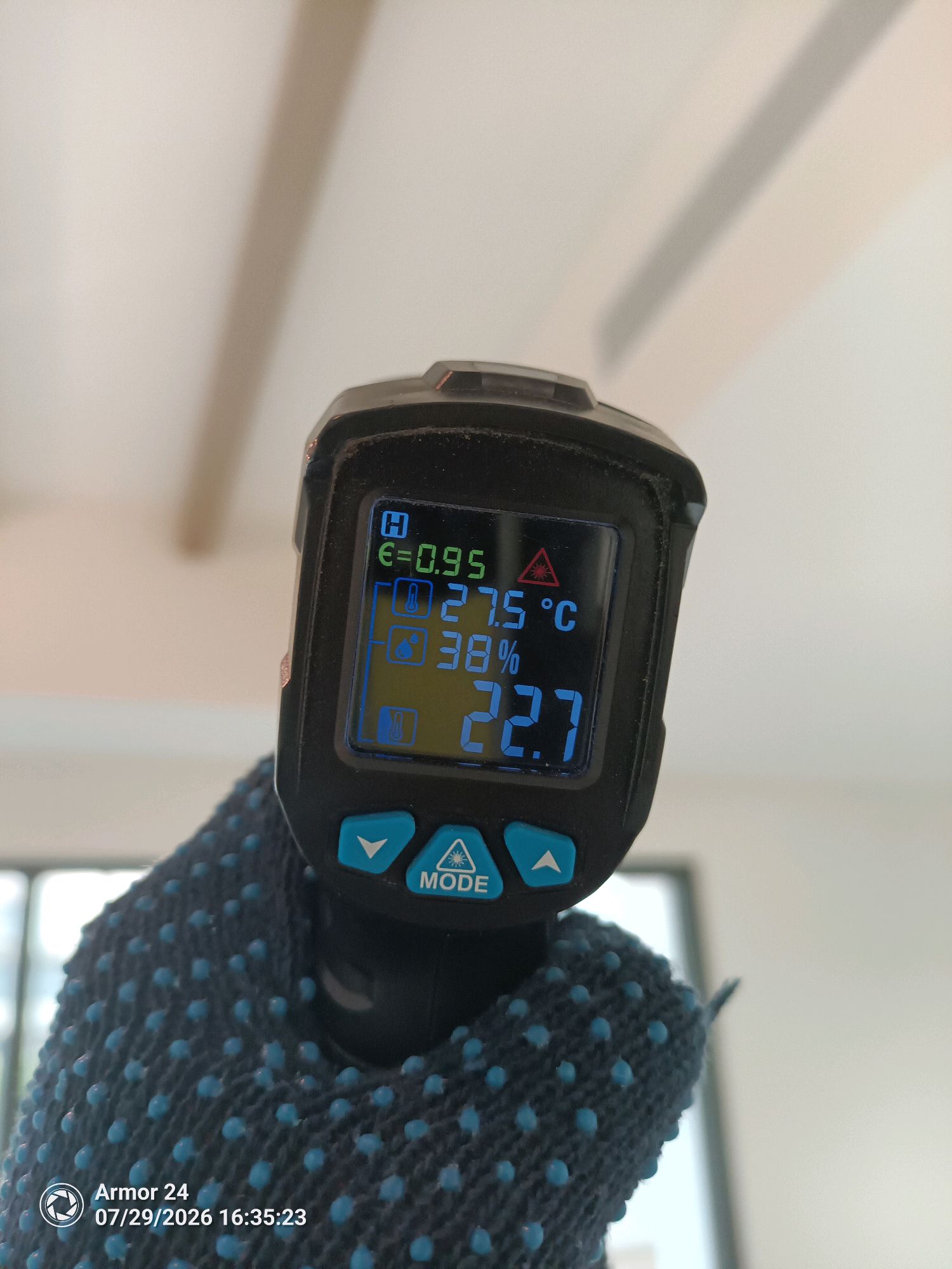 Handheld infrared thermometer displaying 27.5°C and 38% humidity, wrapped in a blue knit sleeve.