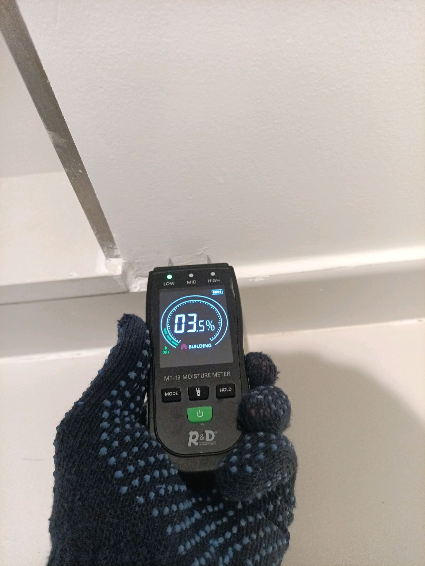 Gloved hand holds a MT-19 moisture meter against a light wall; the device screen shows 03.5% and the word DRY, indicating low moisture.