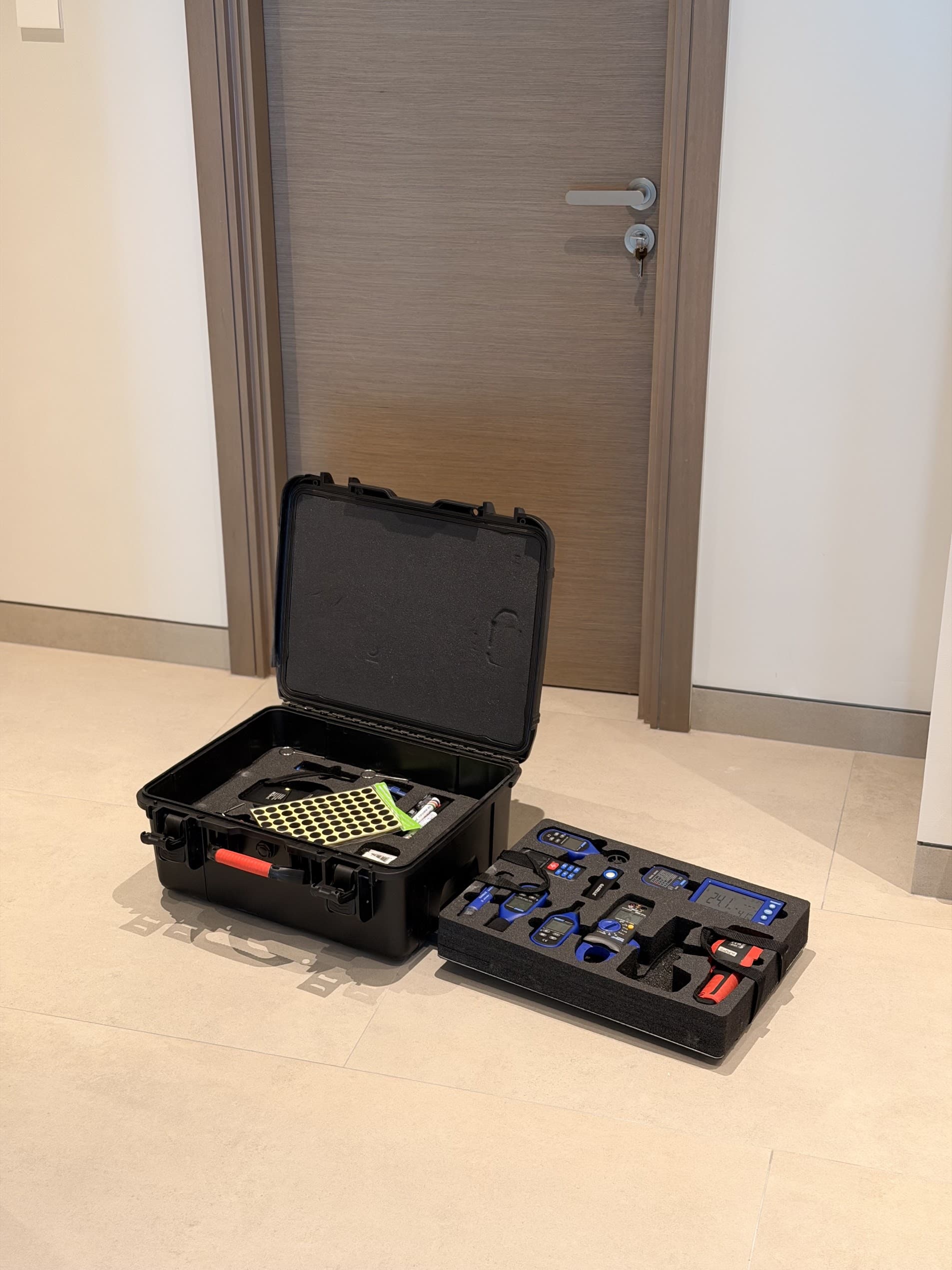 Open black tool case on a light tiled floor with blue-handled tools and a yellow perforated insert, in front of a wooden door.