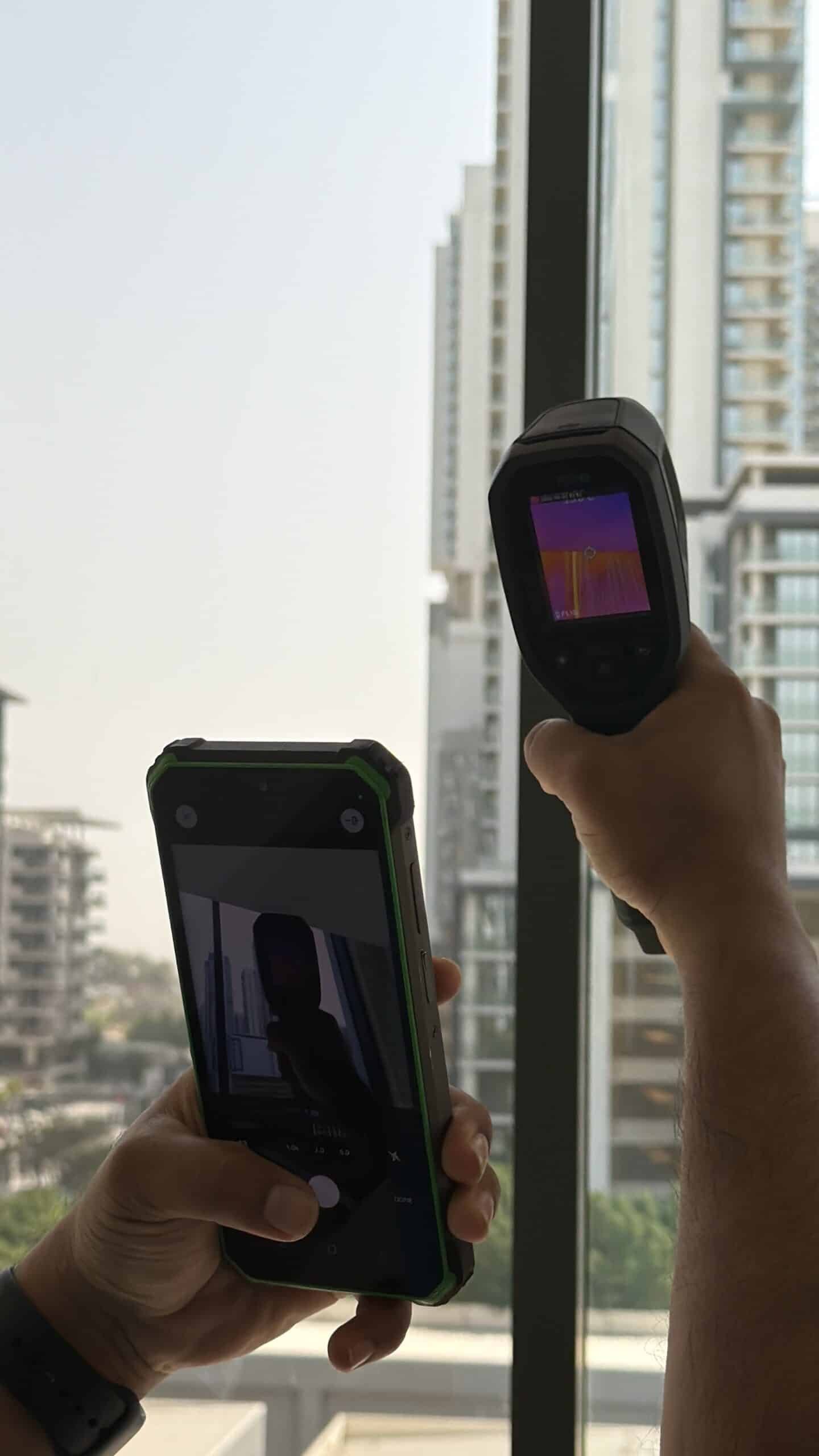 Two hands hold a handheld thermal camera displaying a purple heat-map image, while another hand holds a smartphone to capture the scene by a window with city buildings outside.